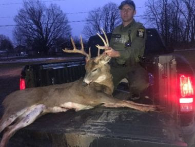 evening photo of ECO holding deceased buck by antlers in back of a pickup truck