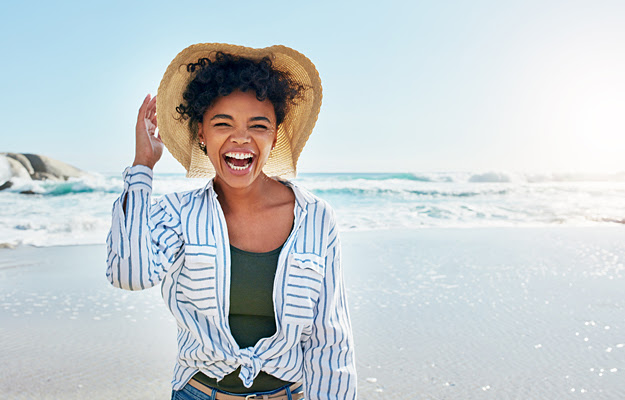 A woman smiling on the beach with a sun hat and long sleeves on.