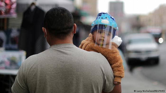 Bebê usando uma proteção de acrílico contra o coronavírus é carregado por um homem nas ruas dos Estados Unidos