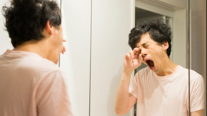 Young man waking up and yawning as he looks in the bathroom mirror.