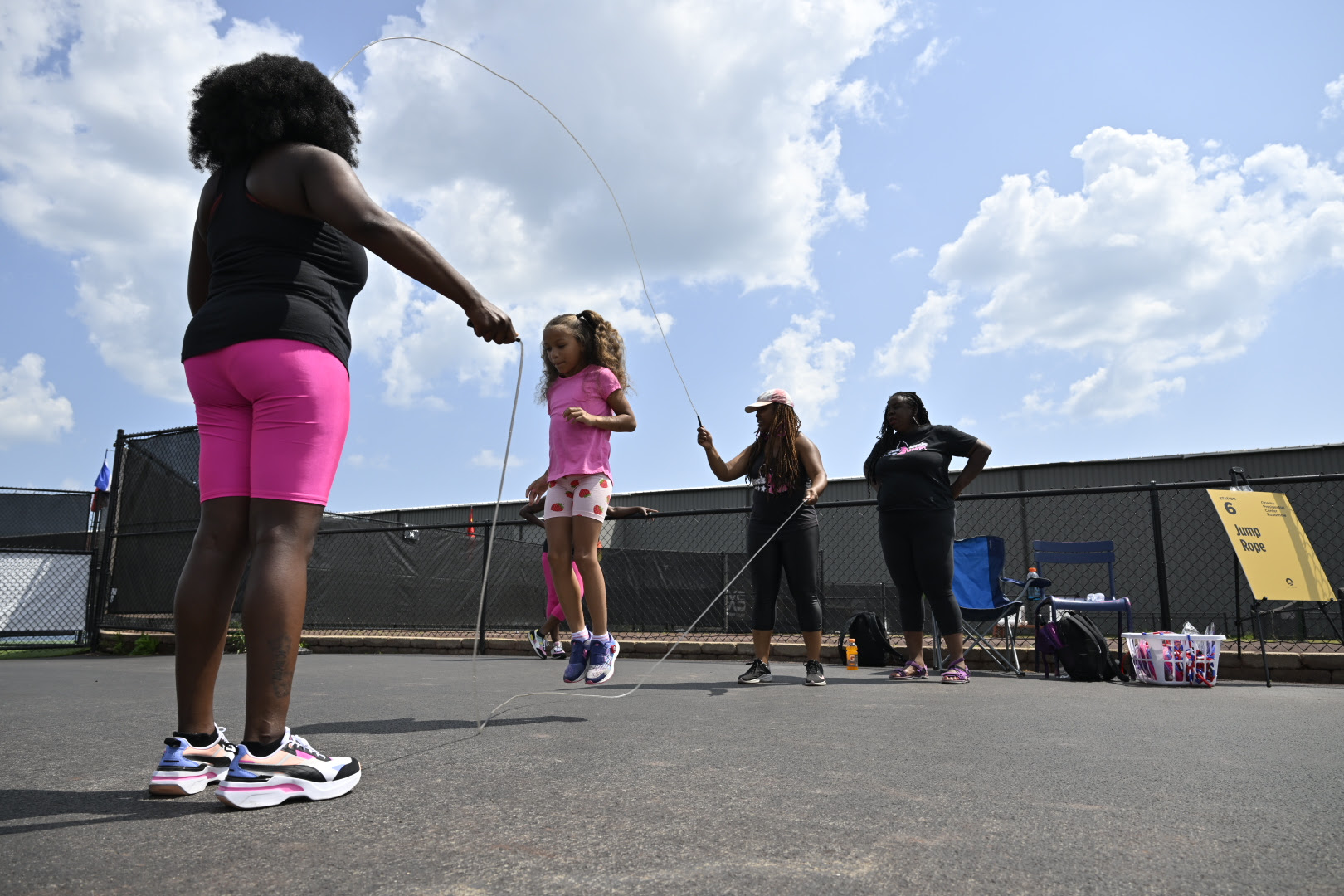 Four people with medium to dark skintones and playing double dutch outside on asphalt.