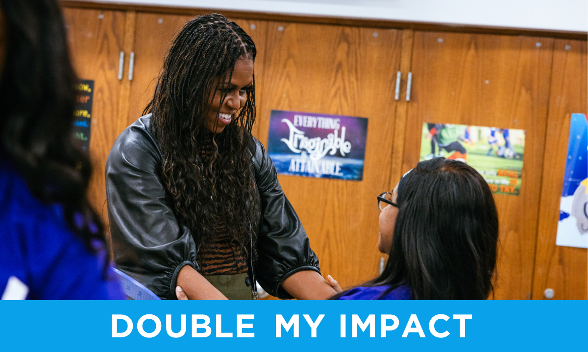 Mrs. Obama wears a black leather jacket and olive green pants. She smiles with both arms outstretched towards a young girl seen in profile, who is wearing a blue shirt, glasses, and a headband on her dark hair. she has medium skin and is holding out both hands in an embrace with Mrs. Obama. A blue bar at the bottom of the image reads "Double my impact" in white text.