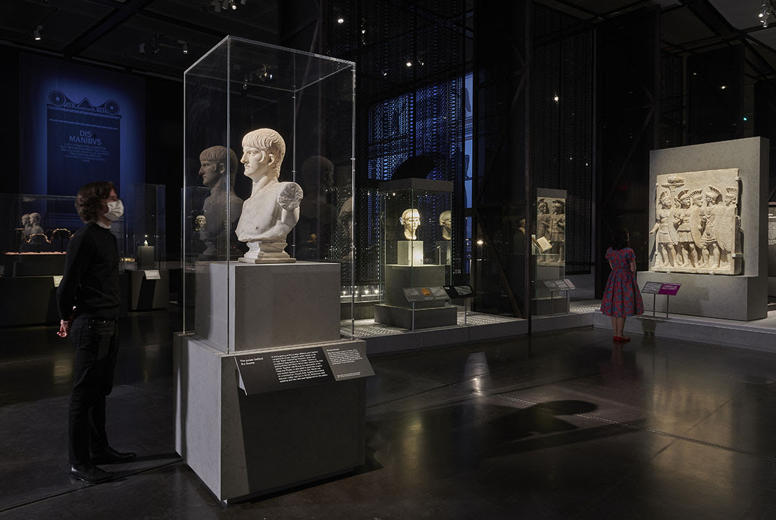 Inside 'Nero: the man behind the myth'. A visitor looks at a marble bust of Nero to the left, and another visitor looks at a sculpture of Roman soldiers in the background. A blue projection can be seen behind.