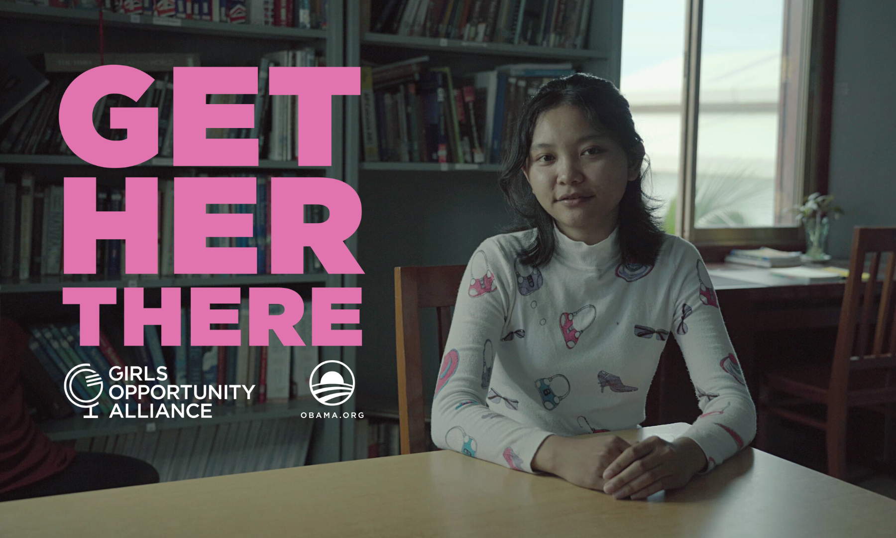A young girl with light skin and dark, shoulder-length hair looks to camera with her arms crossed on the table in front of her. She sits in a classroom with books behind her. To her left, “Get Her There” is featured in pink lettering with the Girls Opportunity Alliance and Obama Foundation logos below.