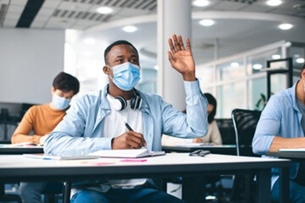 Black male student in mask with hand raised at desk