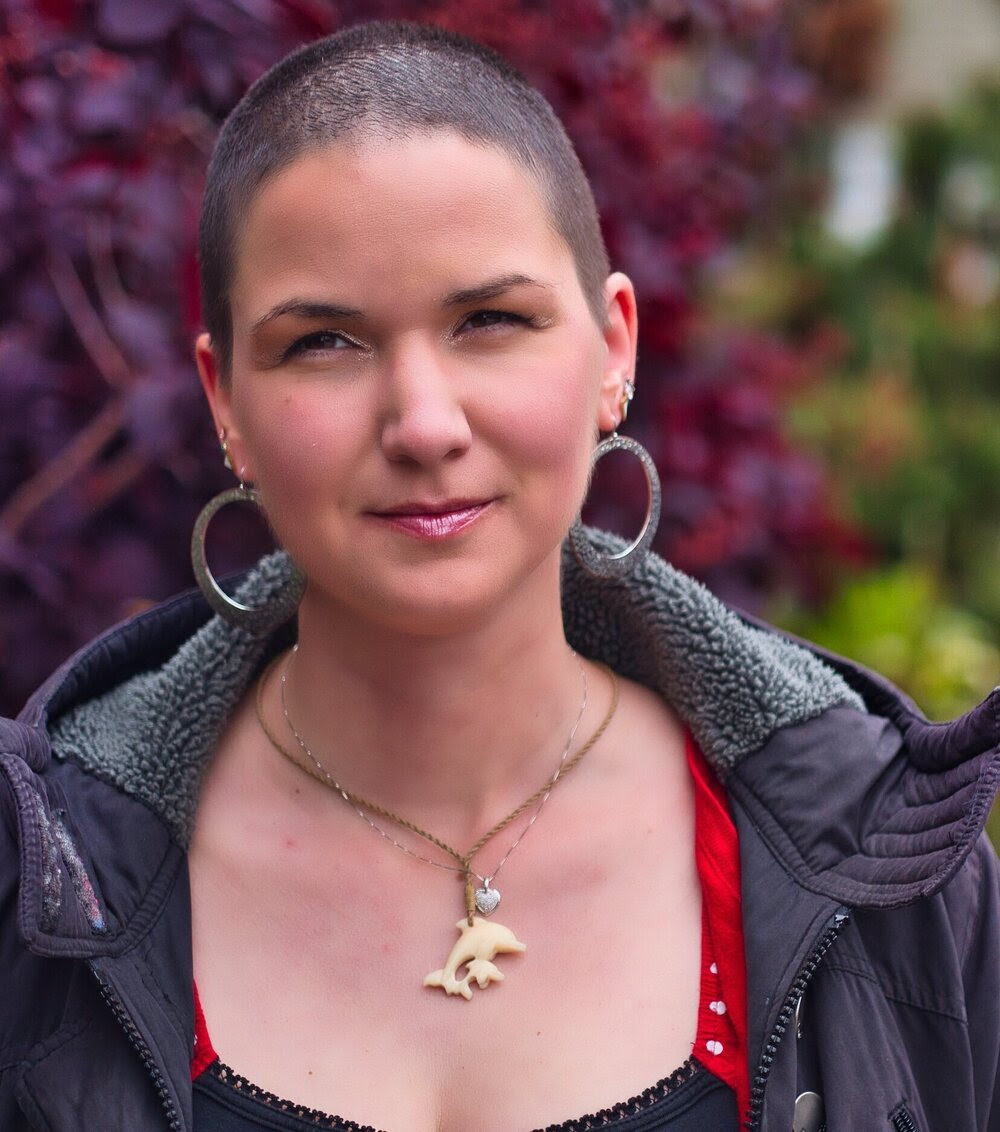 Karina, a woman with super short brown hair. She is wearing a necklace showing two dolphins and large silver earrings.