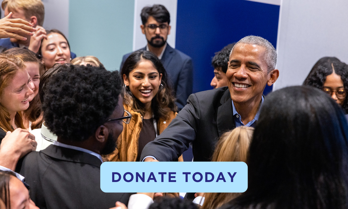 President Obama wears a navy suit jacket and light blue collared shirt, smiling at and shaking hands with a young man with short dark hair and a deep skin tone. Light blue and royal blue panels are in the background, as well as a group of smiling young people of a range of skin tones who are looking on. An aqua button reads “Donate Today” in black text.