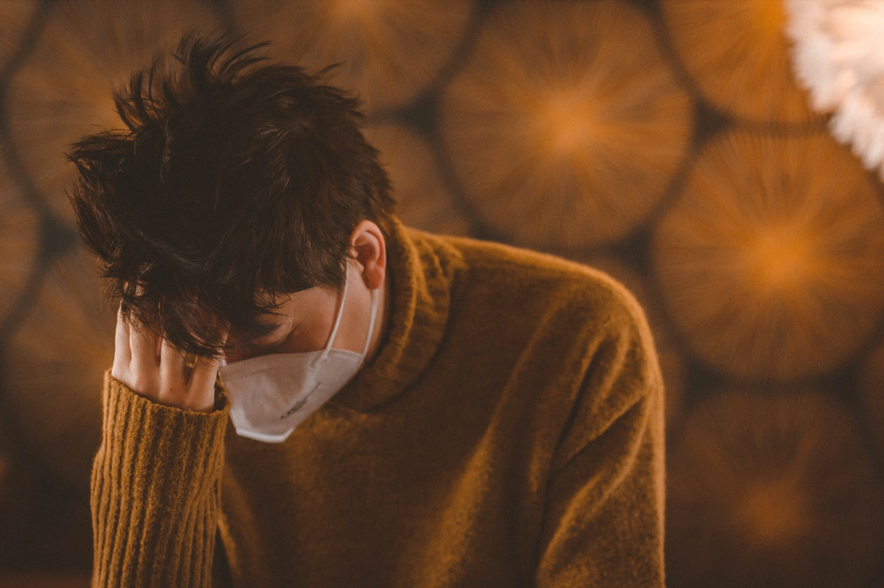 a person wearing a surgical face mask rests their head in their hand looking tired