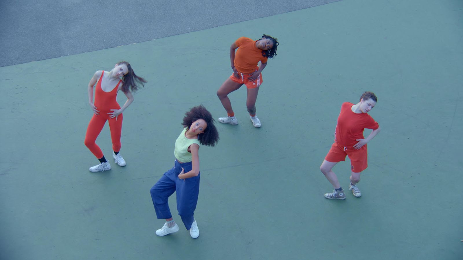 Four dancers are shot from overhead; they are dancing on an empty basketball court, the bright colors of their clothes contrasting with the light green ground. Three of the dancers wear oranges and red, and the dancer in the foreground is wearing yellow and blue. They are bending their legs slightly, hands on front of their hips, with their heads tilted to the left. 