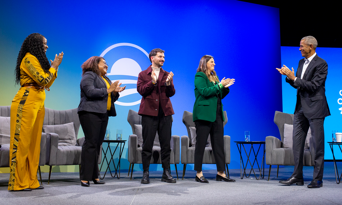 Doussouba Konaté, Landisang Kotaro, Nick Antipov, Natalia Herbst, and President Obama clap as they stand on a stage at the Obama Foundation Democracy Forum. All speakers are of medium and deep skin tones. In the background is a graphic that reads “Democracy Forum.”