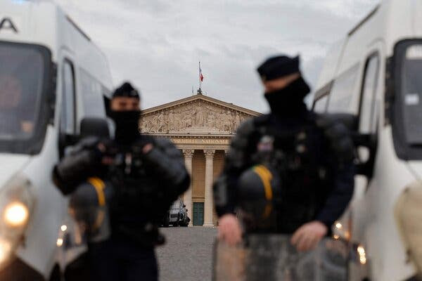 Riot police officers and vans, blurred in the foreground, with the columned National Assembly building clear in the background.