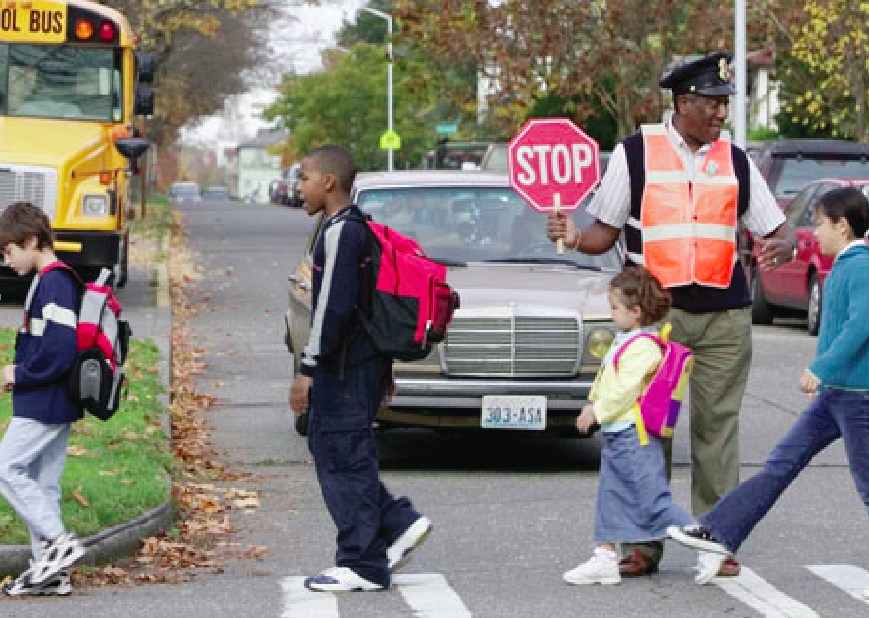 A Black man in an orange vest holds traffic while four children of varying ages cross the street in front of him.