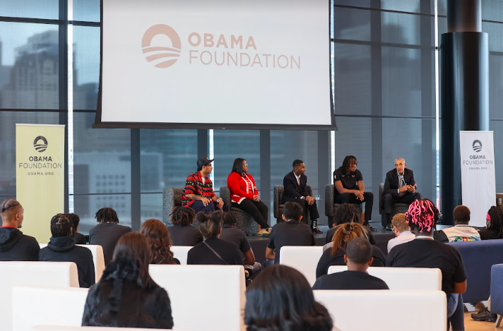 President Obama speaks on a panel alongside Ayo Dosunmu, point guard for the Chicago Bulls, Adeeb Borden, and Aniya Hill, moderated by Chicago influencer and entrepreneur Don. C. The speakers are a range of darker skin tones, and they all sit on a stage facing an audience of people with a range of skin tones. Multiple signs in the background read,“Obama Foundation.”