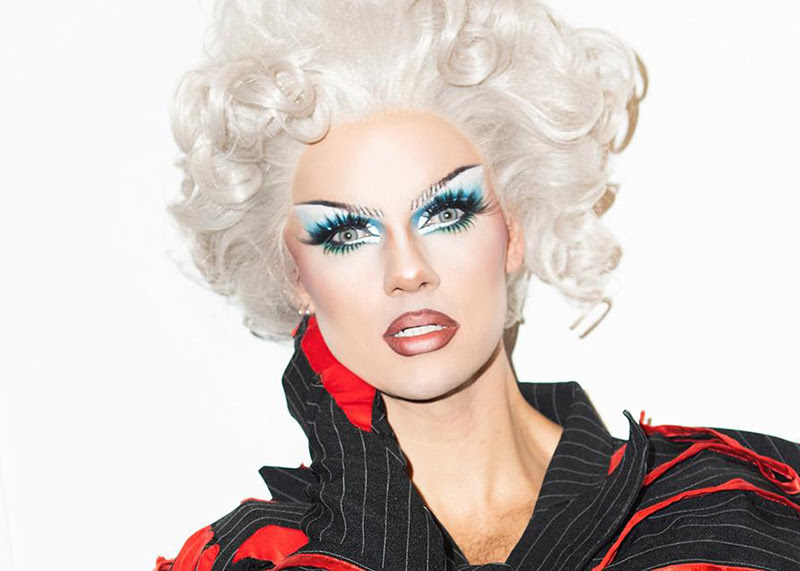A photo of drag artist Crystal. Crystal has white blonde curly hair and a painted face with blue eye shadow.