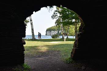 Two people walking with harbor and cliffs in background, seen through brick archway