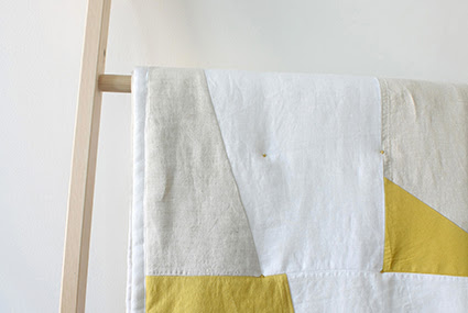 A photograph of a quilt by Woolgathering. It is displayed on a wooden ladder against a white wall, and is made of beige, white and yellow fabric.