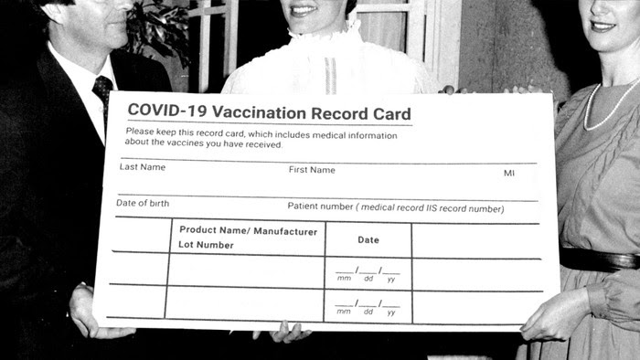 Three well-dressed people hold an enormous, fake COVID-19 vaccination card.
