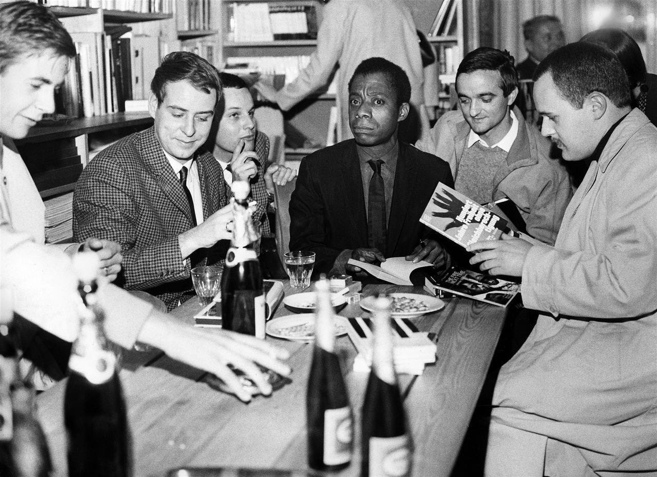 James Baldwin sits at a table surrounded by white men