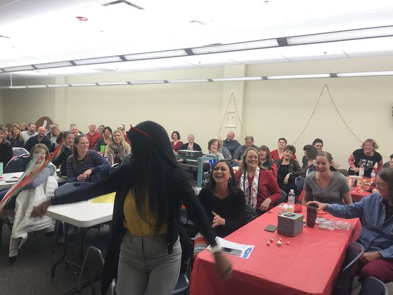 WDE staff sit at tables in a filled conference room for the all-staff meeting while a WDE employee leads a flash mob by waving a red and green ribbon wand in the air.