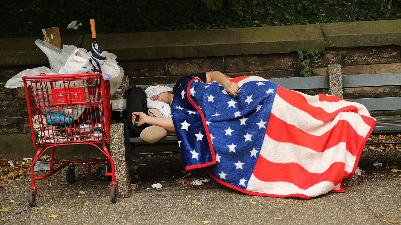 A homeless man sleeps under an American flag blanket on a park bench in New York City. New U.S. data reports a drop in the number of homeless people — but not in New York and other states.