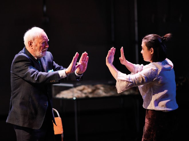 An older person and younger person on stage, with their hands outstretched facing each other