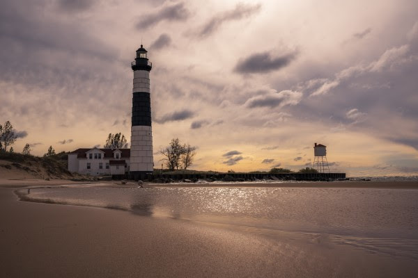 A lighthouse stands tall against the backdrop of purple-gray dusk as gentle waves lap at a sandy shore.