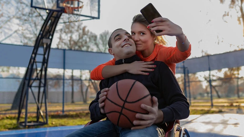 At a basketball court, a woman embraces a man in a wheelchair as they pose for a photograph she is taking using a smartphone.