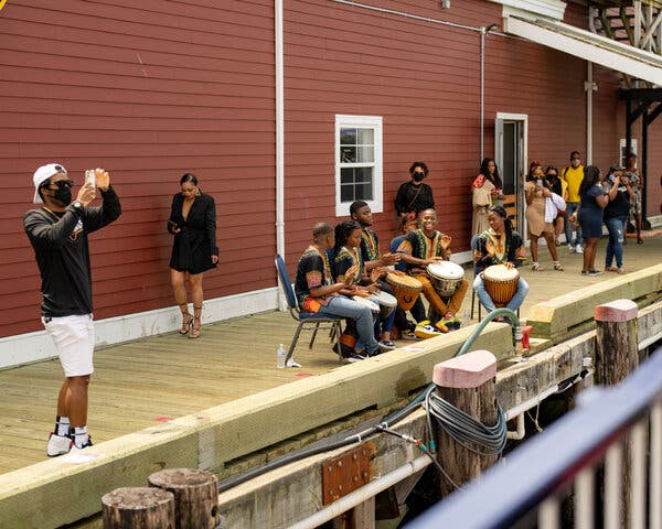 Outside a reddish building on a dock, groups of Black tourists stand, while a group of five drummers in traditional dress, play hand drums.