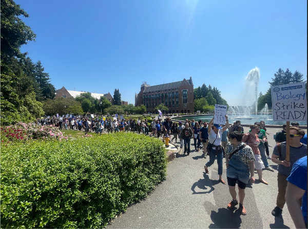 People march past Drumheller fountain. In the foreground is a person with a sign that says "Biology Strike READY," and behind them another person holds a sign that reads "B*TCH Better Have My $$$"