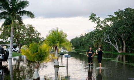 Two people stand and talk in a flooded street surrounded by trees