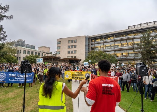 A crowd of 1,000 workers on the UC San Diego campus listening to a speaker. Workers in the front of the crowd are holding a blue UAW 5810 banner, a yellow Student Researchers United banner, and a red UAW 2865 banner.