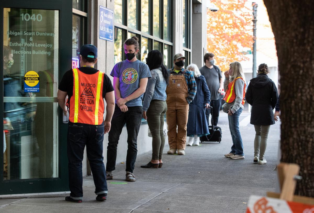 Voters stand in line outside the Multnomah County Duniway-Lovejoy Elections Building. Two election workers help in orange vests.
