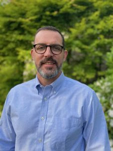 Tom Flanagan stands smiling, wearing a blue button-down shirt and glasses, in front of a tree