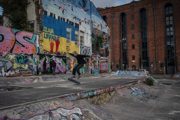 A young man is airborne while skating on a concrete expanse near a graffitied wall and a brick building.