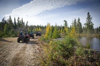 three off-road vehicles travel single file down a dirt road, alongside a large blue body of water. Green forests and blue sky line the background