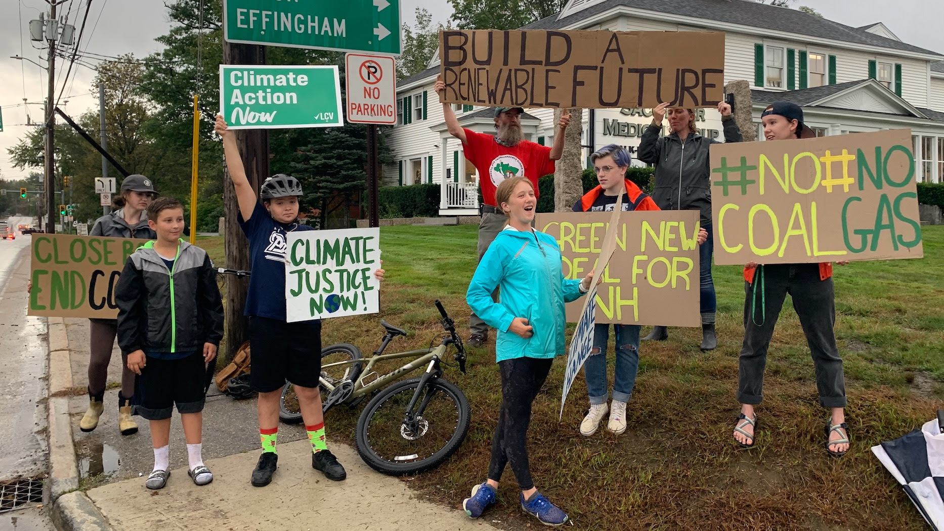 group of young people holding various cardboard signs that say No Coal No Gas, Green New Deal for NH, Build a Renewable Future, Climate Justice Now. They're in raincoats standing on a corner of a street.