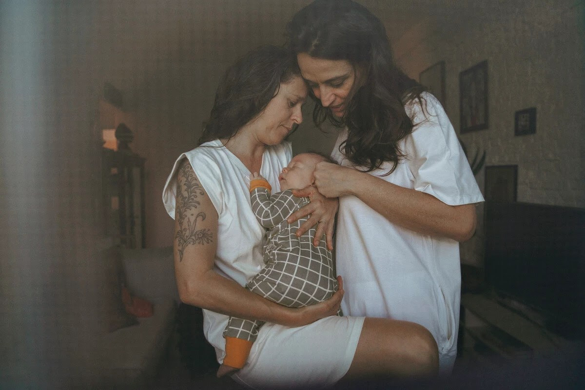 Juliana, Milene e Dom durante período de licença-maternidade. Na imagem vemos o casal abraçado com o bebê Dom no colo. 