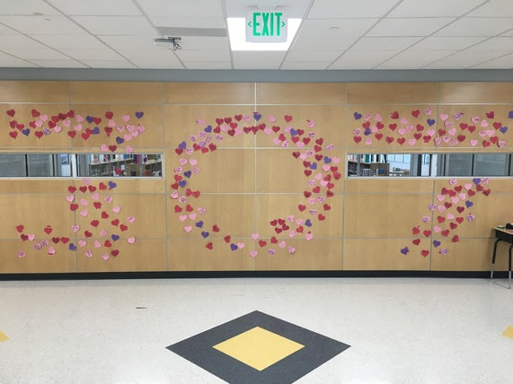 Pink, red, and purple heart-shaped sticky notes are posted on a school wall in a shape that reads "307". Each of the notes has had descriptions of random acts of kindness written on them.