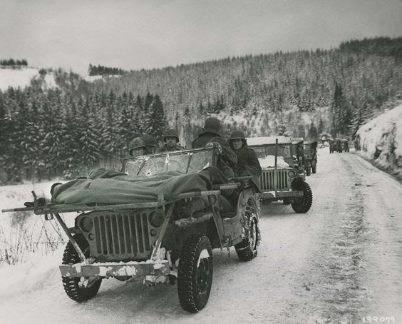 Bullet holes in the windshield of this Jeep are a          testament to the faithful service of the Medics of the 84th          Division as they evacuate wounded soldiers in their makeshift          Jeep ambulance. The Medics are carrying two soldiers wrapped          up in blankets to keep them warm in the frigid cold. The          litters are strapped to a makeshift frame attached to the          flat-hooded Jeep 4x4. This Jeep caravan was part of the 1st          Battalion, 334th Infantry Regiment. Image was taken on January          9th, 1945. Thanks to...