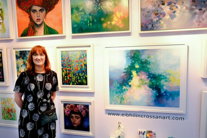 Artist Eibhilin Crossan, in a black dress, stands in front of a number of paintings