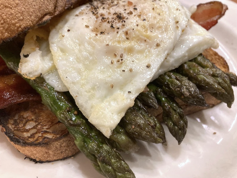Close-up photo of toast with bacon, asparagus, and a fried egg on top.