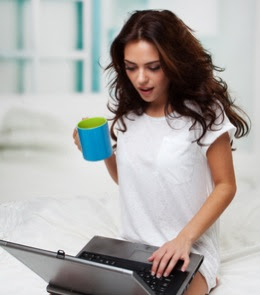 woman using a laptop sitting on bed and drinking coffee