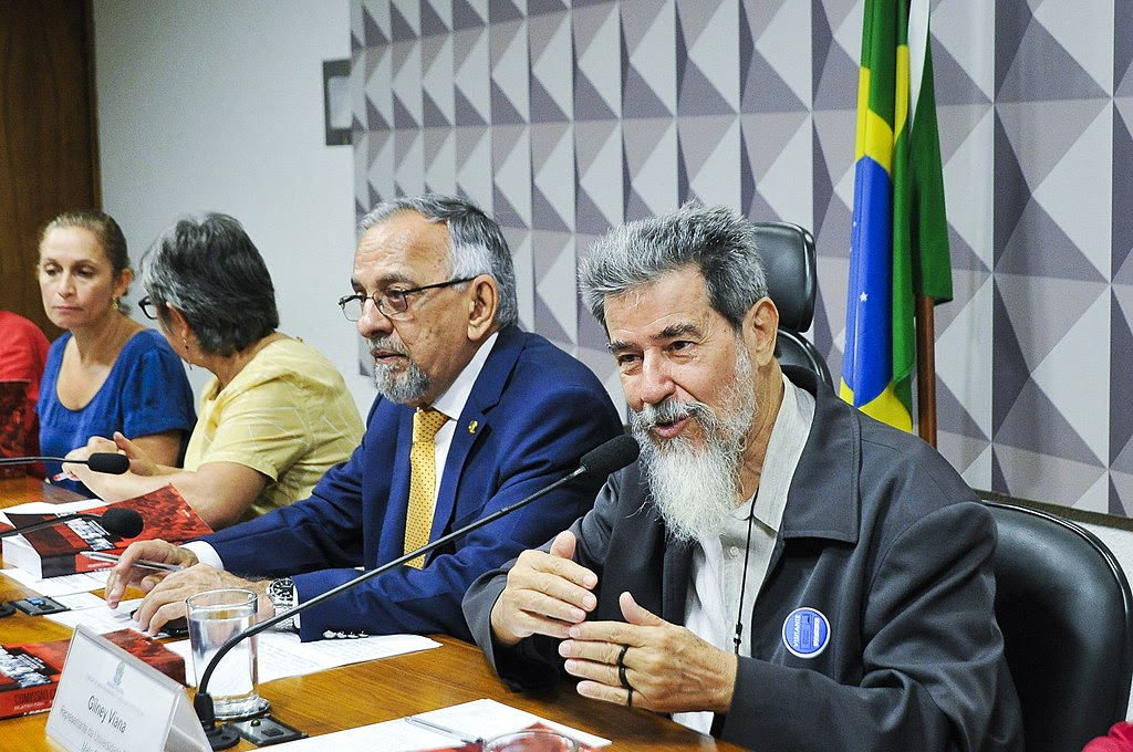 Gilney Viana, ex-preso político da ditadura militar brasileira, é um senhor na faixa dos 70 anos, branco, com cabelos, barba e bigode grisalhos. Ele veste terno cinza escuro e camisa social cinza clara. Gilney está sentado em uma mesa durante uma audiência no Senado relacionado à ditadura militar
