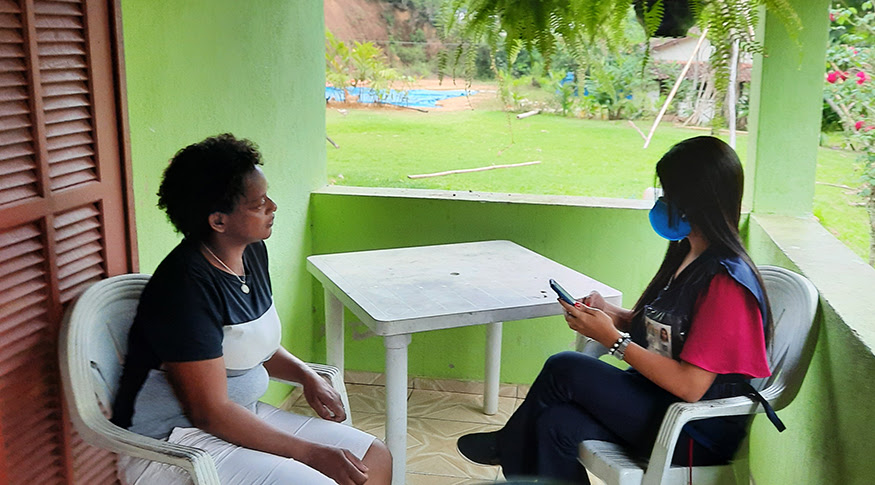 #PraCegoVer A foto mostra a varanda da casa de um quilombo, em que uma moradora (do lado esquerdo) conversa com uma recenseadora uniformizada do IBGE (do lado direito), ambas sentadas em cadeiras brancas com uma mesa branca entre as duas.