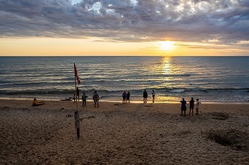several people outlined in shadow stand on a sandy beach during a golden sunset, as waves roll in. A red beach warning flag is posted