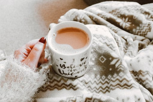 A mug of tea sat on a woman's lap with a grey fleece blanket on her knees viewed from above