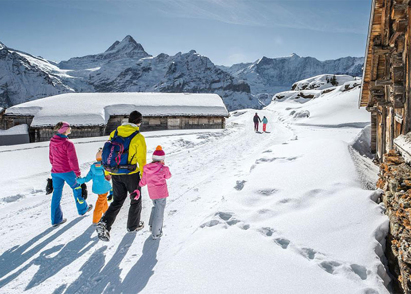 A photo of the Bern region in Switzerland. A family - two parents and two children - walk across the snow in warm jackets and hats. In the background are mountains and blue skies.