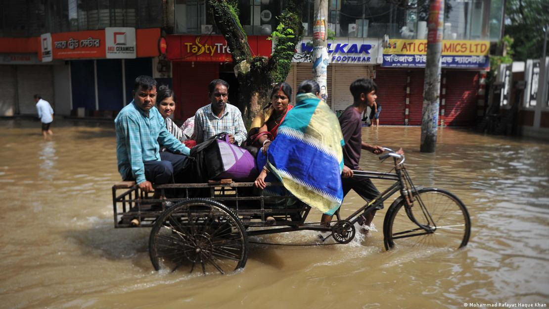 Pessoas sobre carrinho puxado por bicicleta, em meio a enchente em Bangladesh
