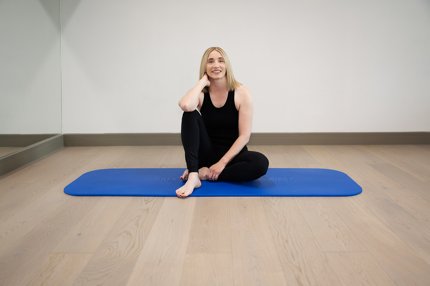 A woman with long blonde hair and a black workout outfit sits on a blue yoga mat. 