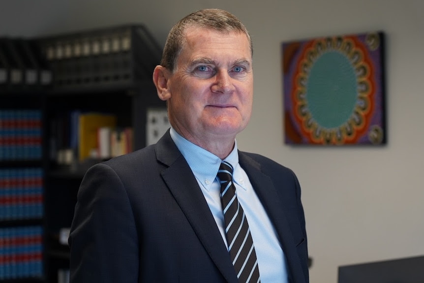 A slightly smiling middle -aged man, short dark blond hair, wearing a suit and tie stands in front of a book shelf, painting.